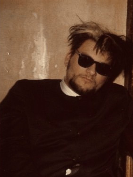 Erick Grau in the 1990s, bleached hair, dark sunglasses and a black suit, slouched against a bar